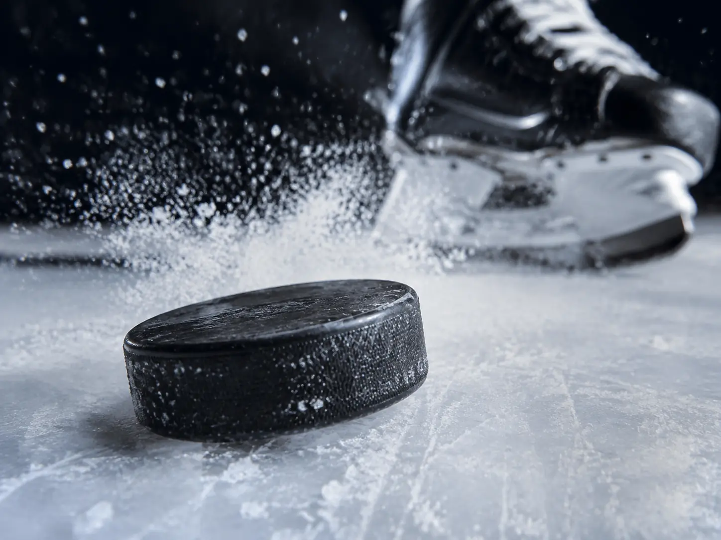 Black hockey puck on ice beside a player’s skates and stick