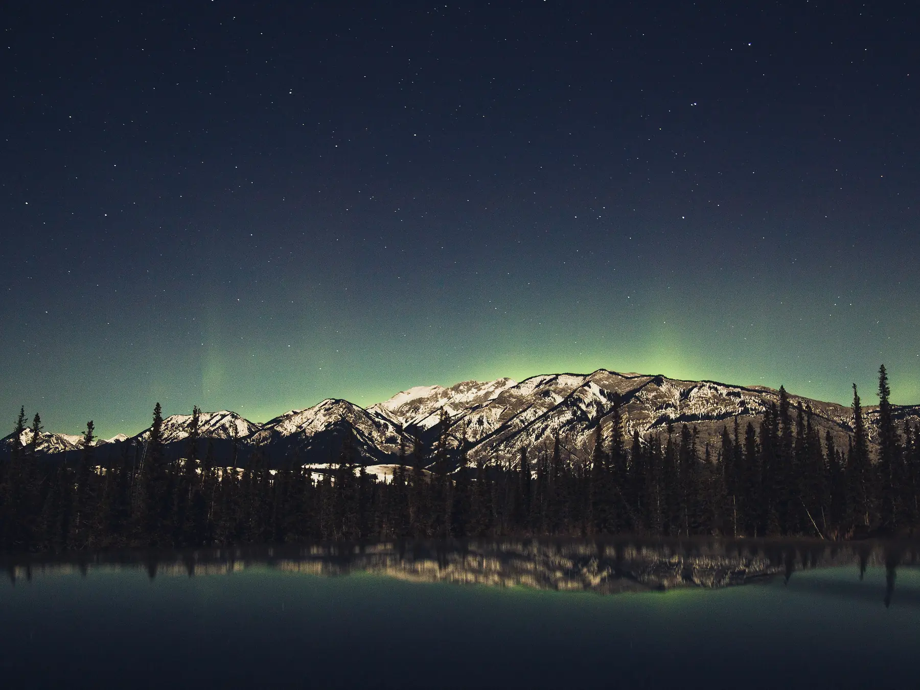 Green northern lights reflected in a calm lake beneath mountains