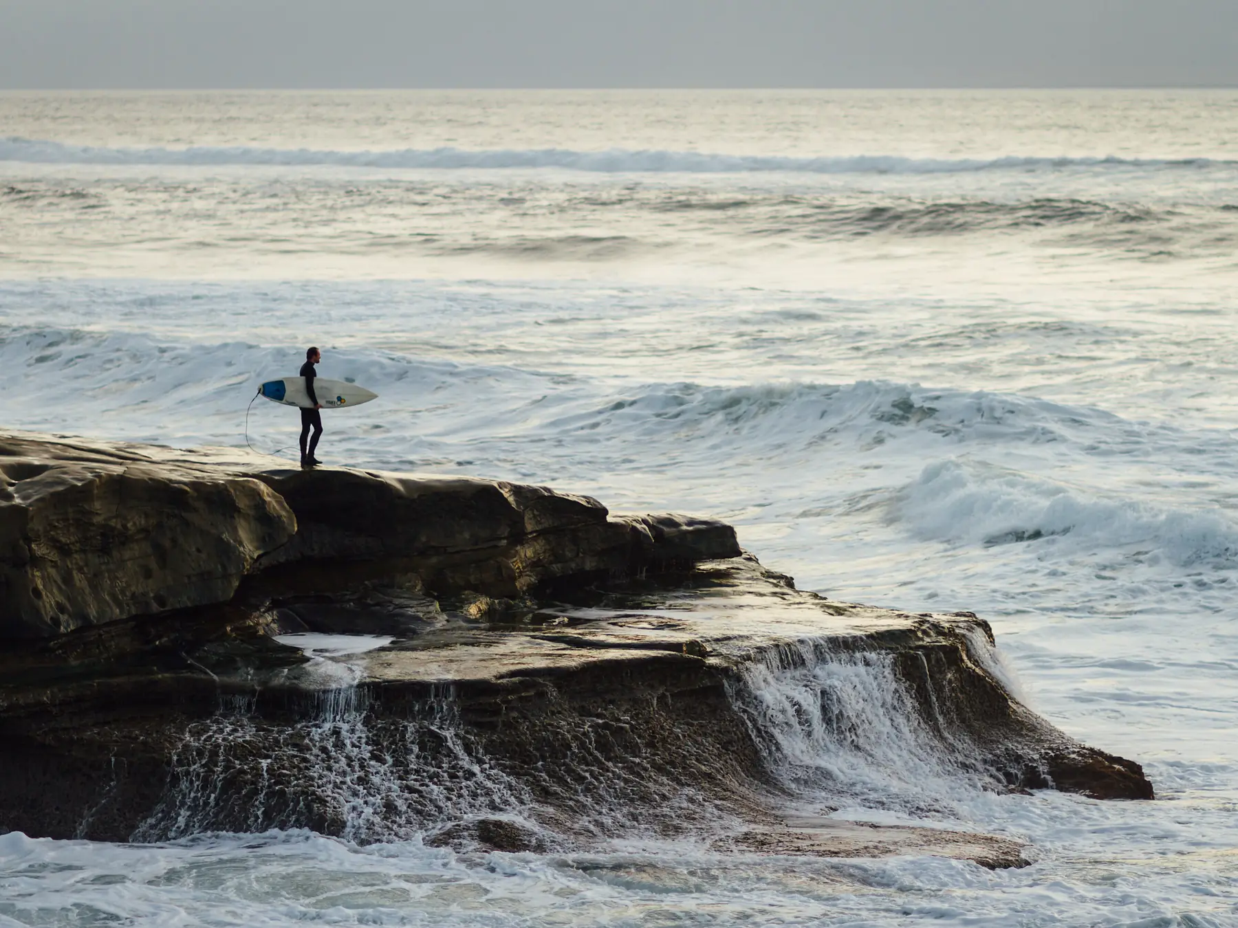 Surfer holding a board while watching waves from a rocky shore