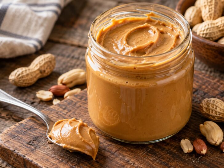 Open jar of smooth peanut butter with spoon and peanuts on rustic table, no label